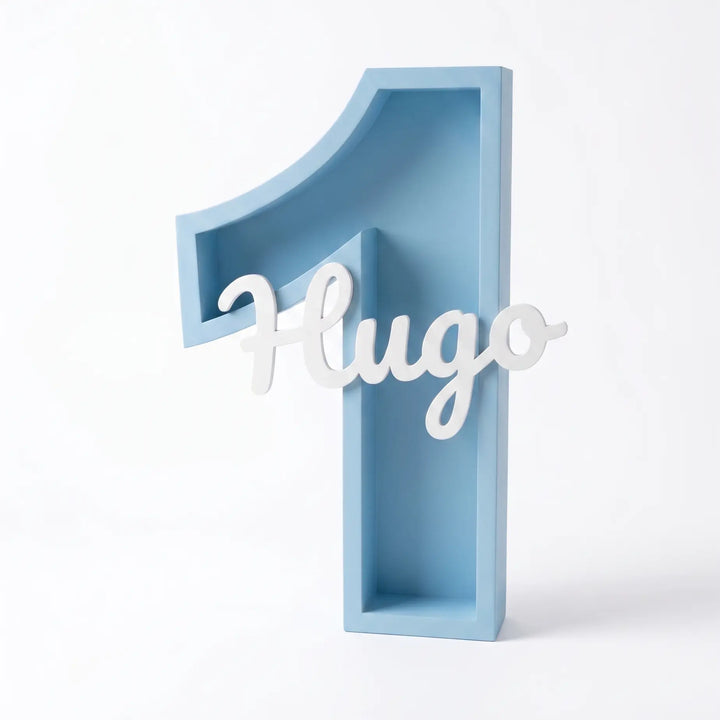 Número 1 decorativo en azul con el nombre Hugo en blanco junto a la tarta de un primer cumpleaños