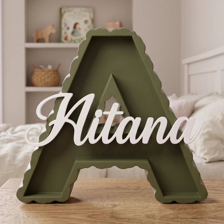 Letra A en verde oliva con el nombre Aitana sobre una mesa junto a la cama en una habitación infantil