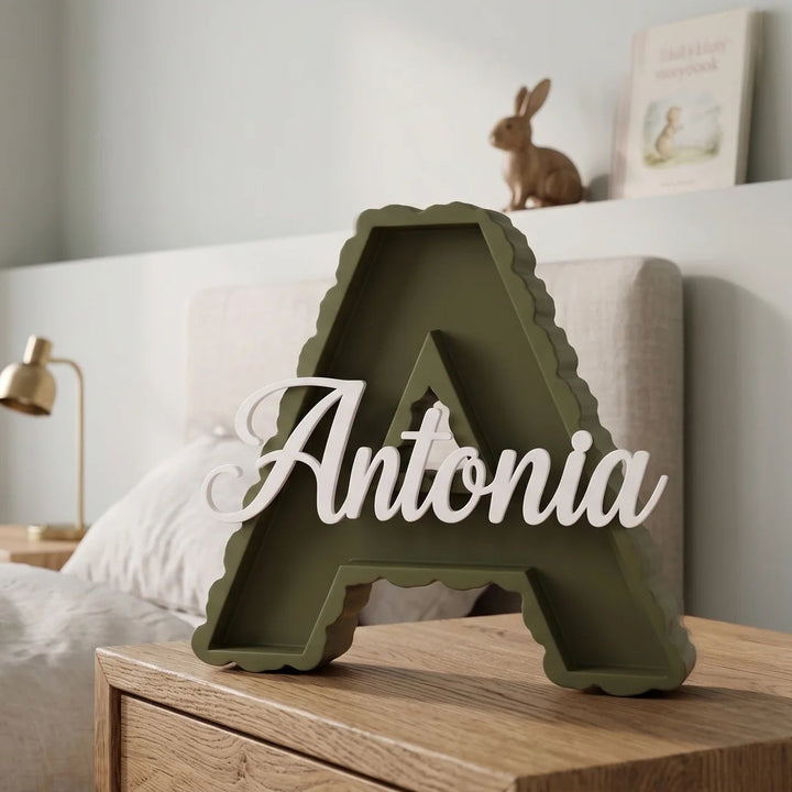 Letra A en verde oliva con el nombre Antonia sobre una mesilla junto a la cama de un dormitorio infantil