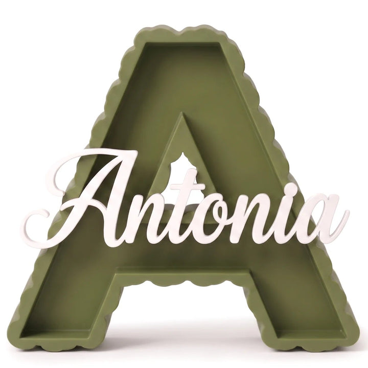 Letra A decorativa de contorno festoneado en verde oliva con el nombre Antonia en caligrafía blanca