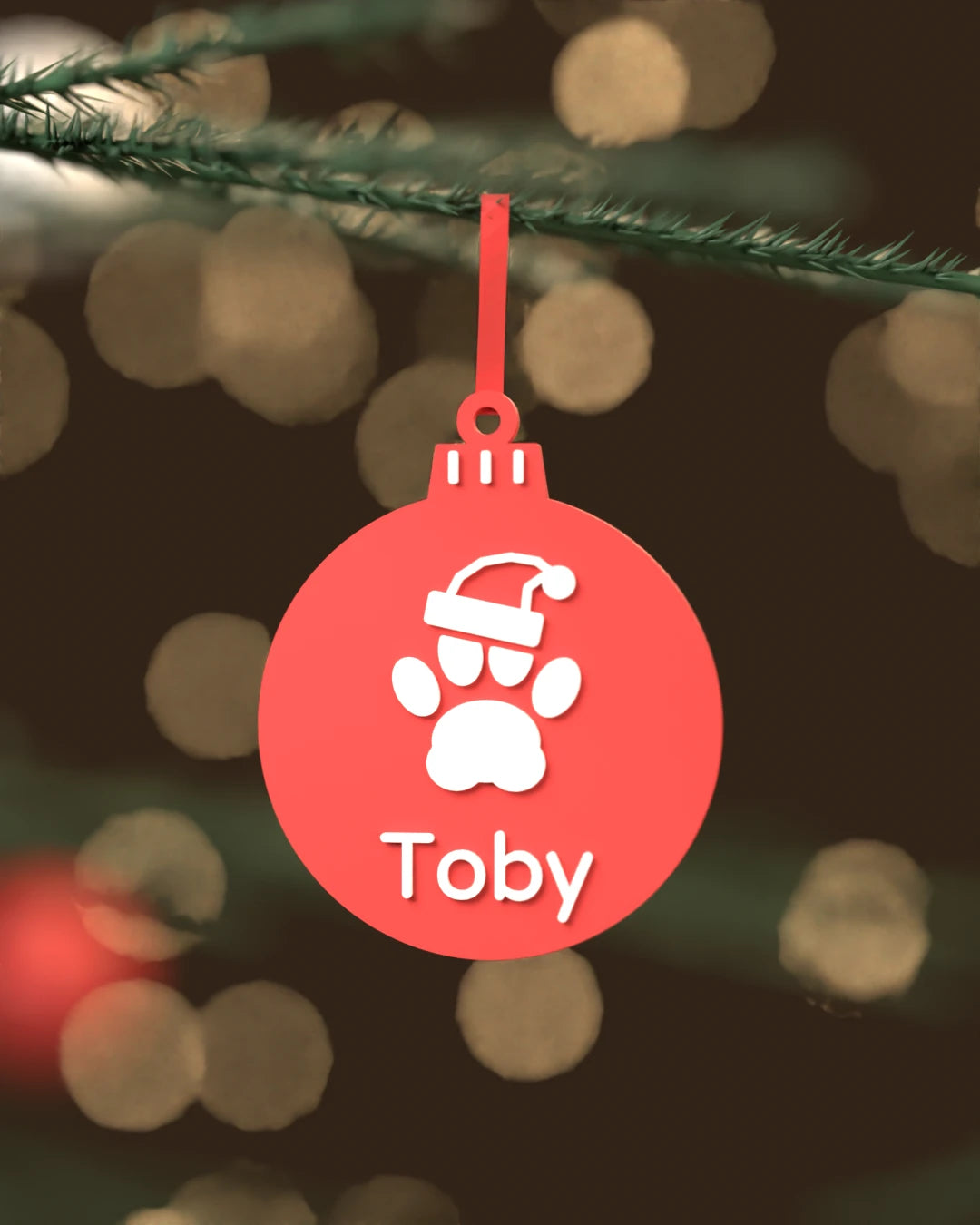 Bola de Navidad Mascota roja con huella, gorro navideño y el nombre Toby en blanco, colgada de una rama de abeto