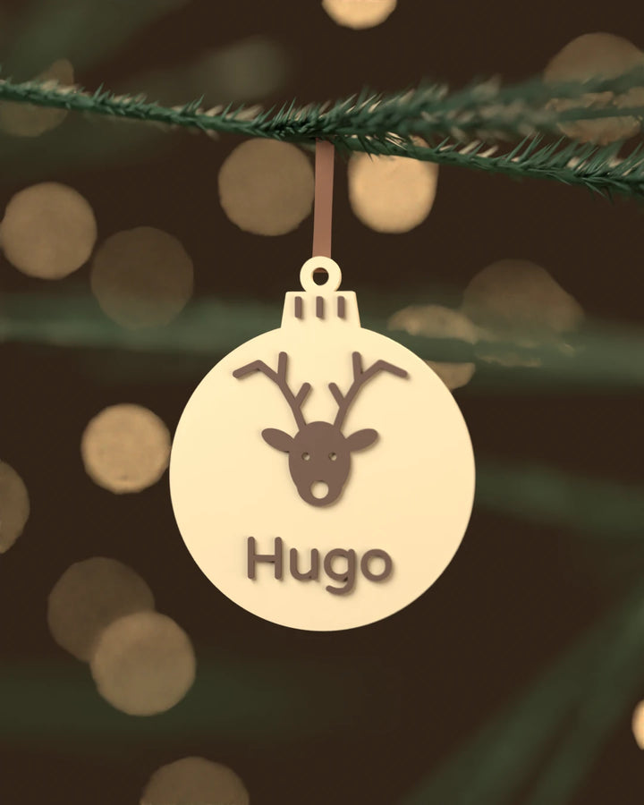 Bola de Navidad Reno color blanco hueso con el nombre Hugo en chocolate oscuro, colgada de una rama de abeto