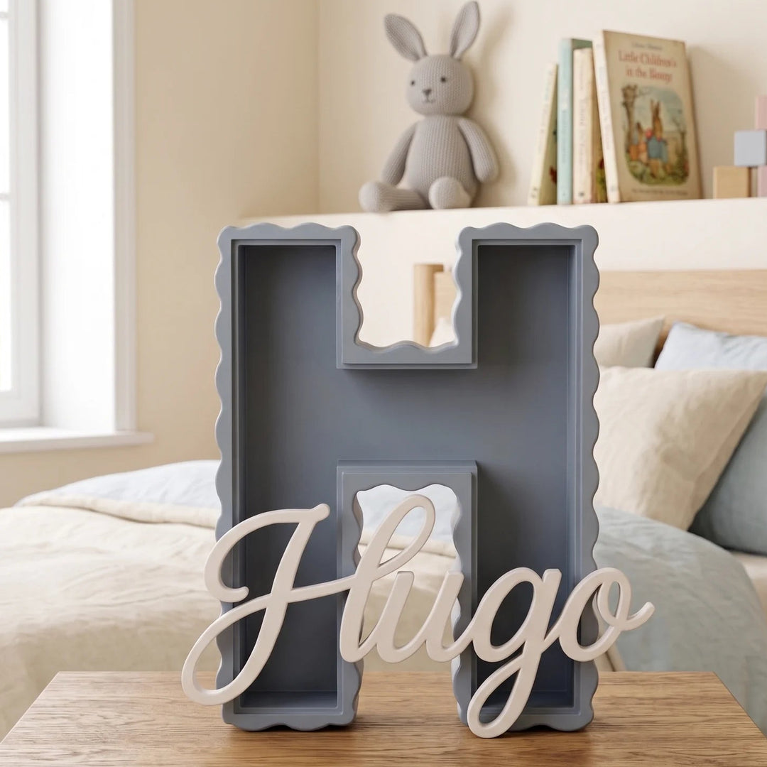 Letra H personalizada 3D de borde ondulado en azul gris con el nombre Hugo en caligrafía sobre una mesita