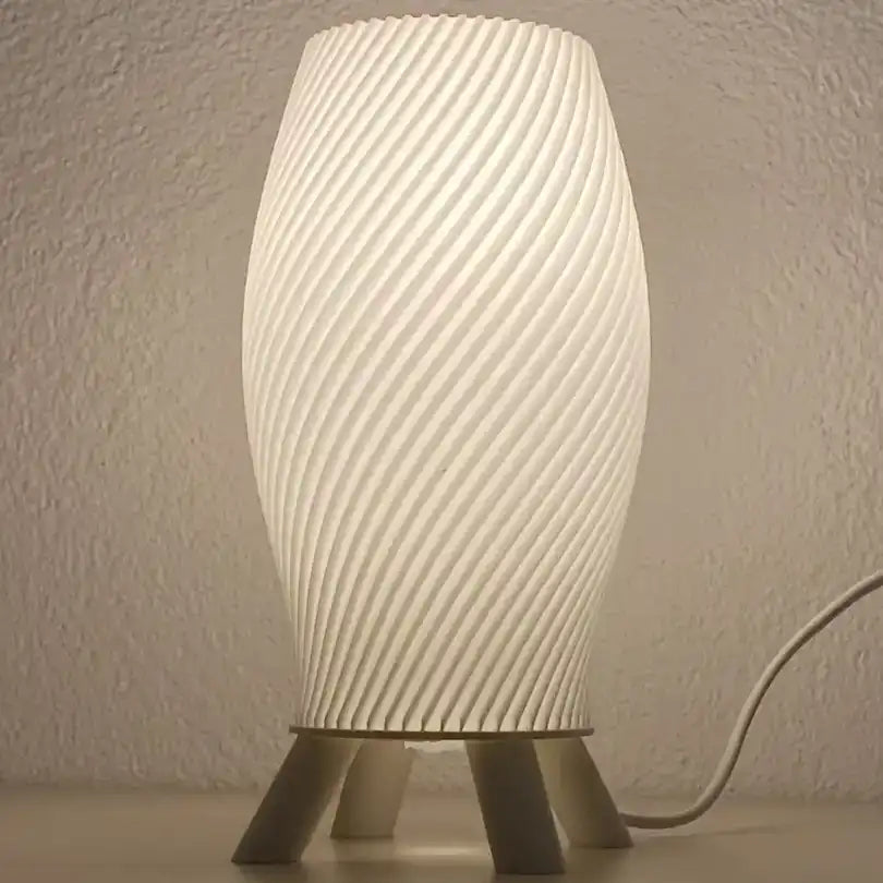 Lámpara de mesa de diseño helicoidal en color crema con base trípode de patas oscuras efecto madera sobre superficie blanca.