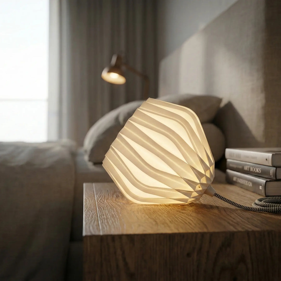 Lámpara de mesa moderna con pantalla blanca facetada y cable textil gris encendida sobre mesita de noche.