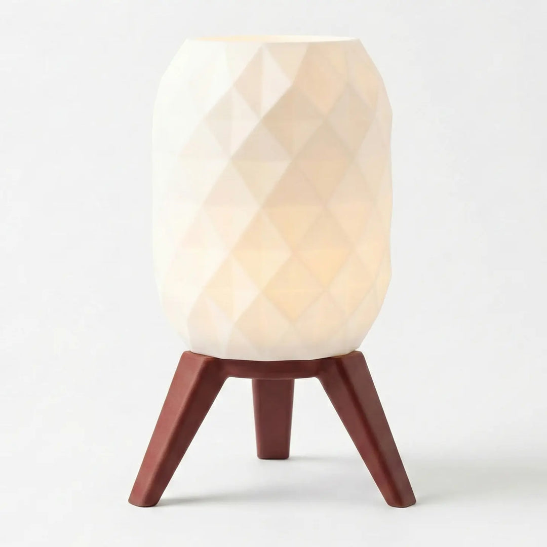 Lámpara de mesa encendida con pantalla geométrica blanca y patas efecto madera clara sobre mesa gris.