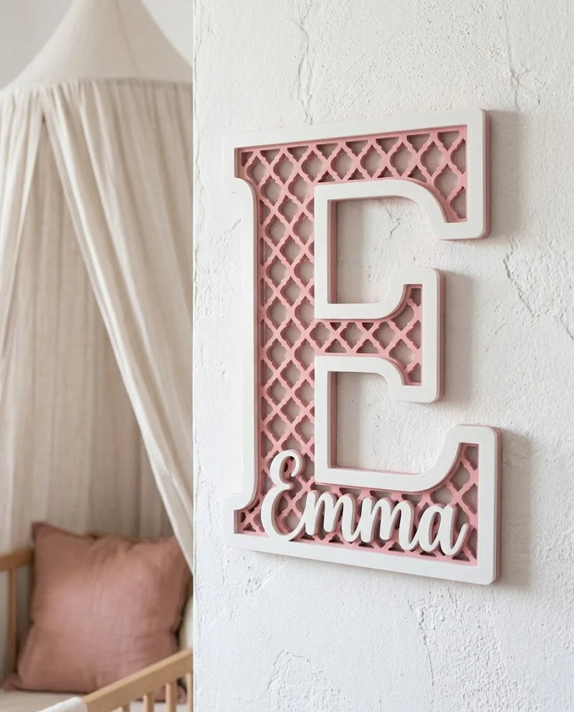 Letra E con el nombre Emma, celosía marroquí calada en rosa sakura y marco blanco, colgada en una habitación de niña mediterránea con dosel de gasa