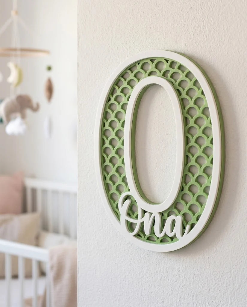 Letra O con el nombre Ona, patrón calado de escamas en verde manzana y marco blanco, colgada en una habitación de bebé en tonos pastel junto a una cuna blanca