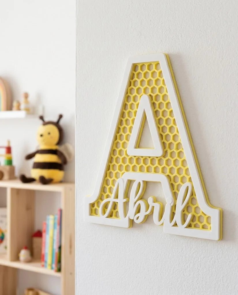 Letra A con el nombre Abril, panal calado amarillo limón y marco blanco, colgada en una habitación infantil luminosa con estantería de madera y peluche de abeja